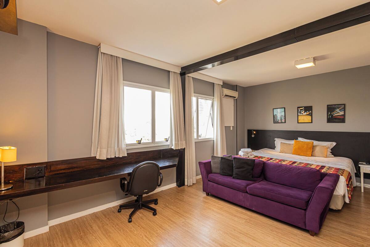 Une chambre d’hôtel avec un lit, un canapé violet, un bureau avec une chaise et une fenêtre avec des rideaux.