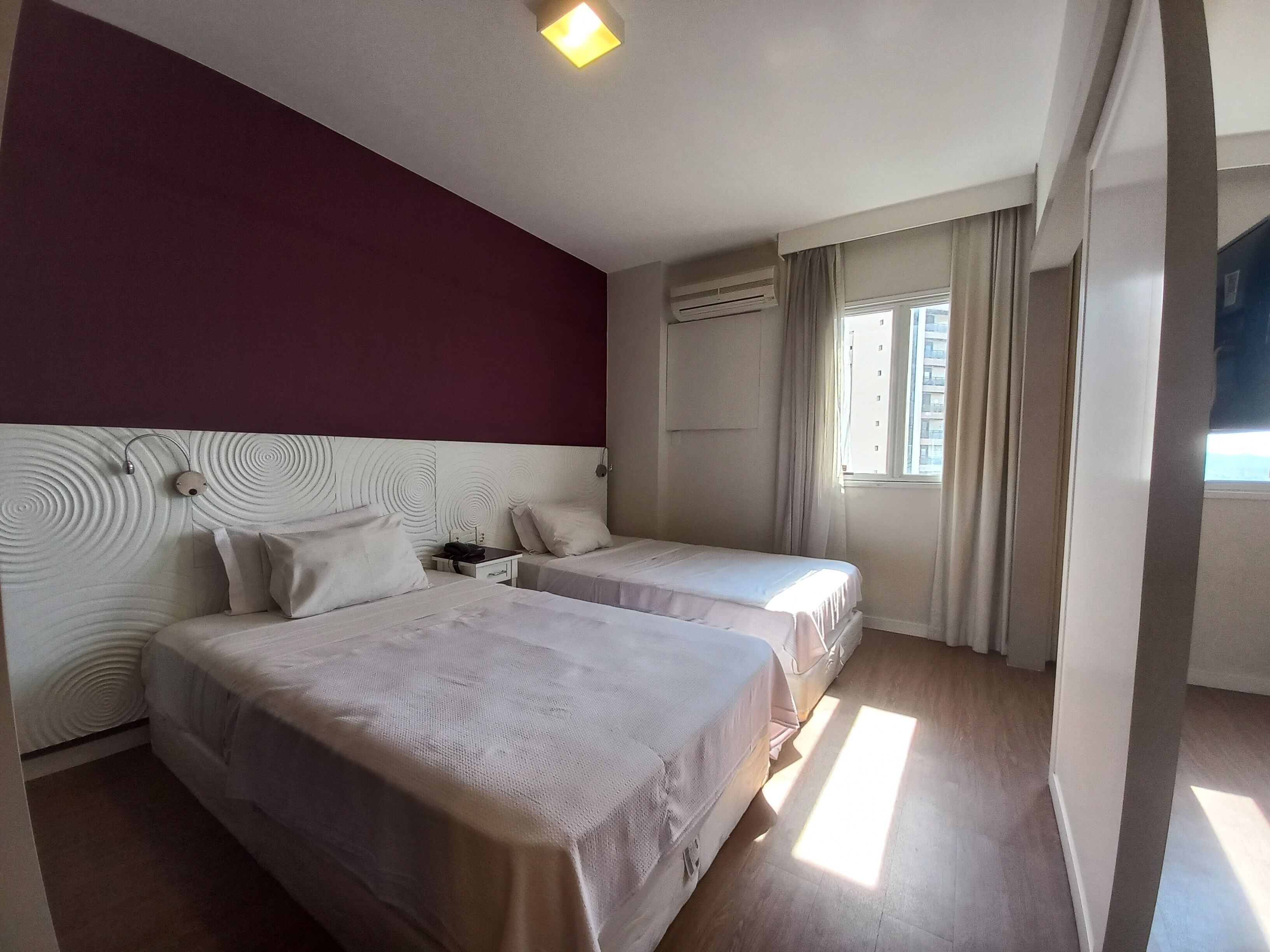 A hotel room with two beds, a red accent wall, a ceiling light, an air conditioning unit, and a window with curtains.