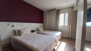 A hotel room with two beds, a red accent wall, a ceiling light, an air conditioning unit, and a window with curtains.