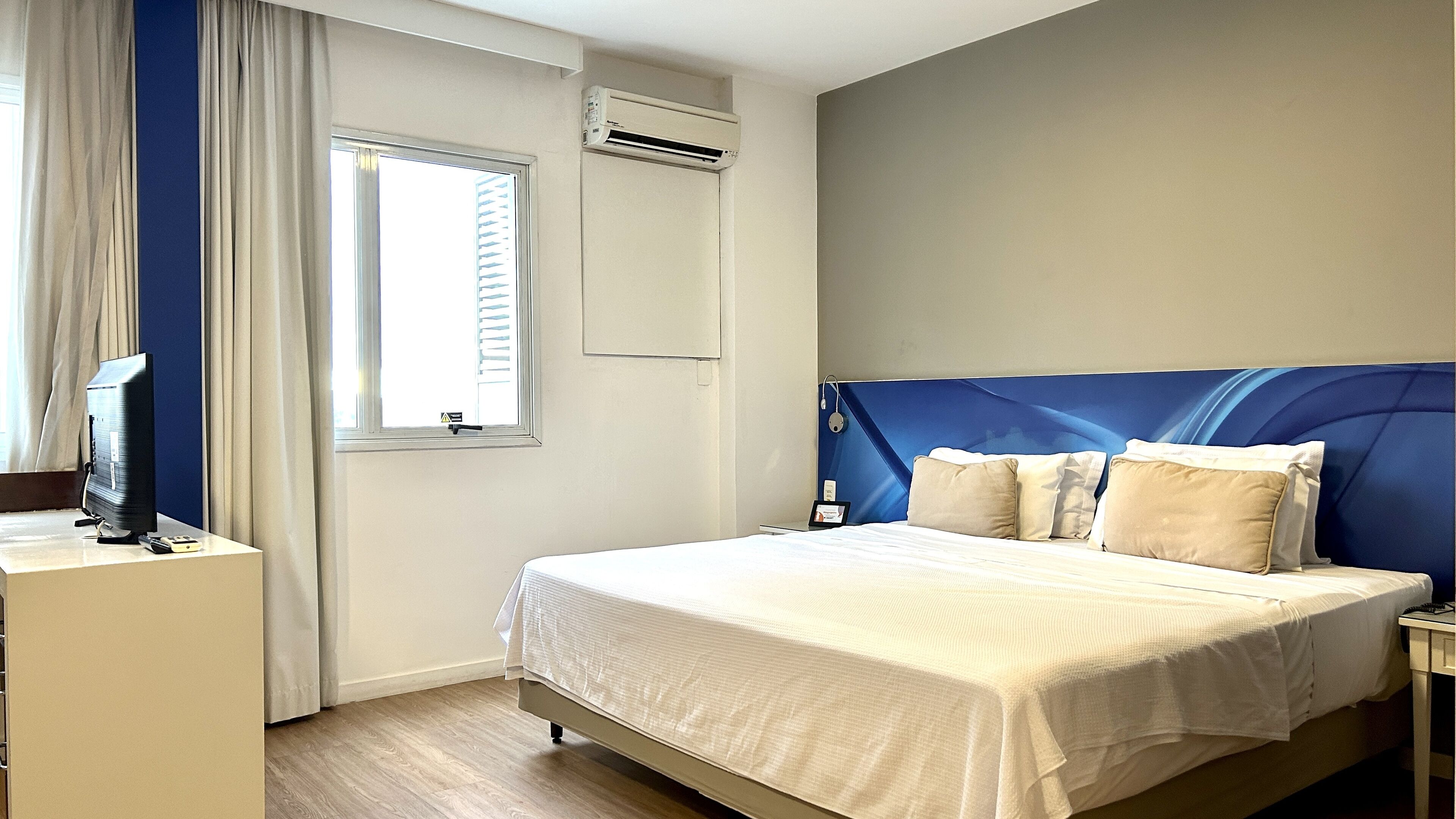 A hotel room with a bed, a desk with a television, and an air conditioning unit.