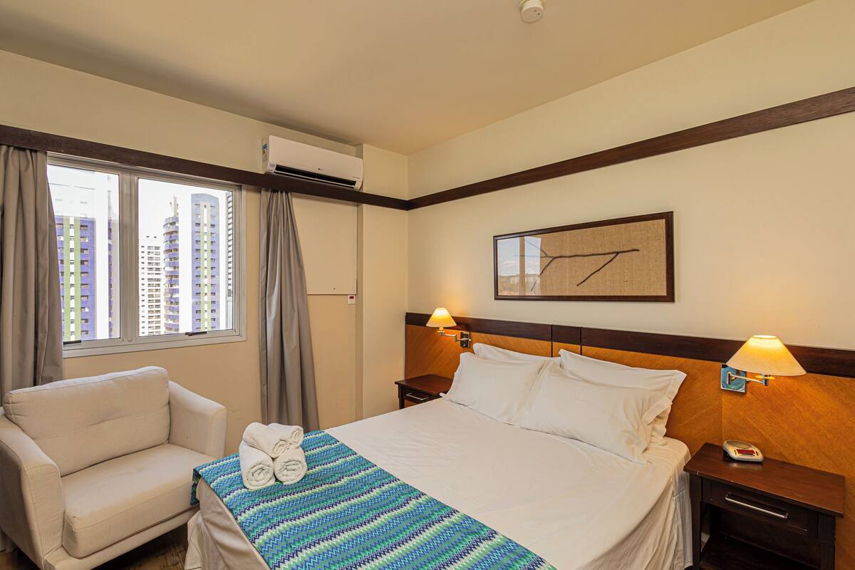 Une chambre d’hôtel comprenant un lit, une chaise, une fenêtre avec des rideaux, un appareil de climatisation, une horloge murale et un tableau encadré.