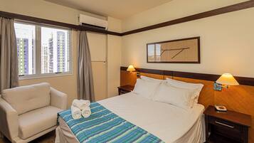 Quarto de hotel com cama, poltrona, janela com cortinas, aparelho de ar condicionado, relógio fixado na parede e quadro emoldurado.