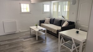 A modern living room with a grey sofa, a white coffee table, and a white side table. There are two windows providing natural light, and a door with a mirror. - Cosy apartment in a 2** residence (Martigues)