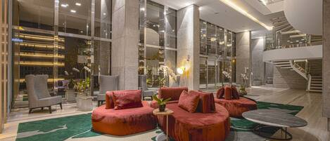 A modern lobby with a seating area featuring red sofas and a round table, large glass windows, and a staircase.