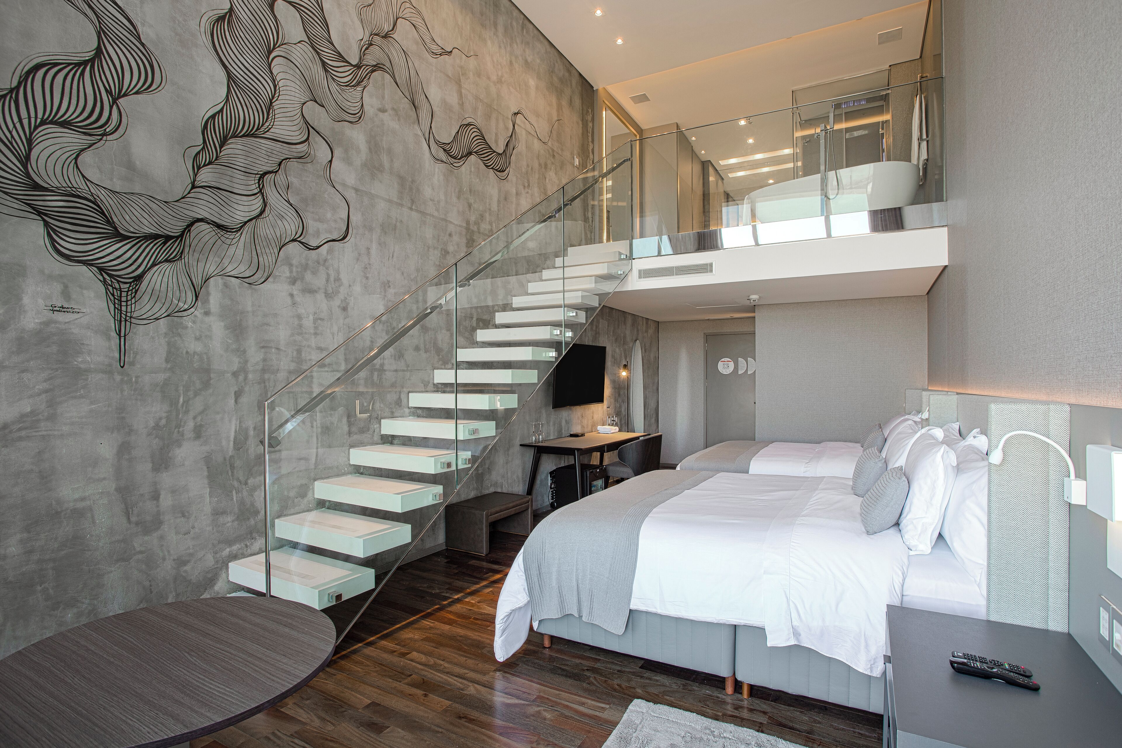 A modern hotel room with a large bed, a desk with a computer, a glass staircase, and a wall art piece.