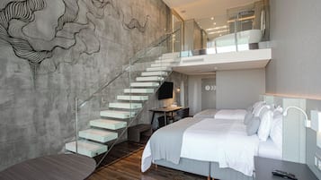 A modern hotel room with a large bed, a desk with a computer, a glass staircase, and a wall art piece.