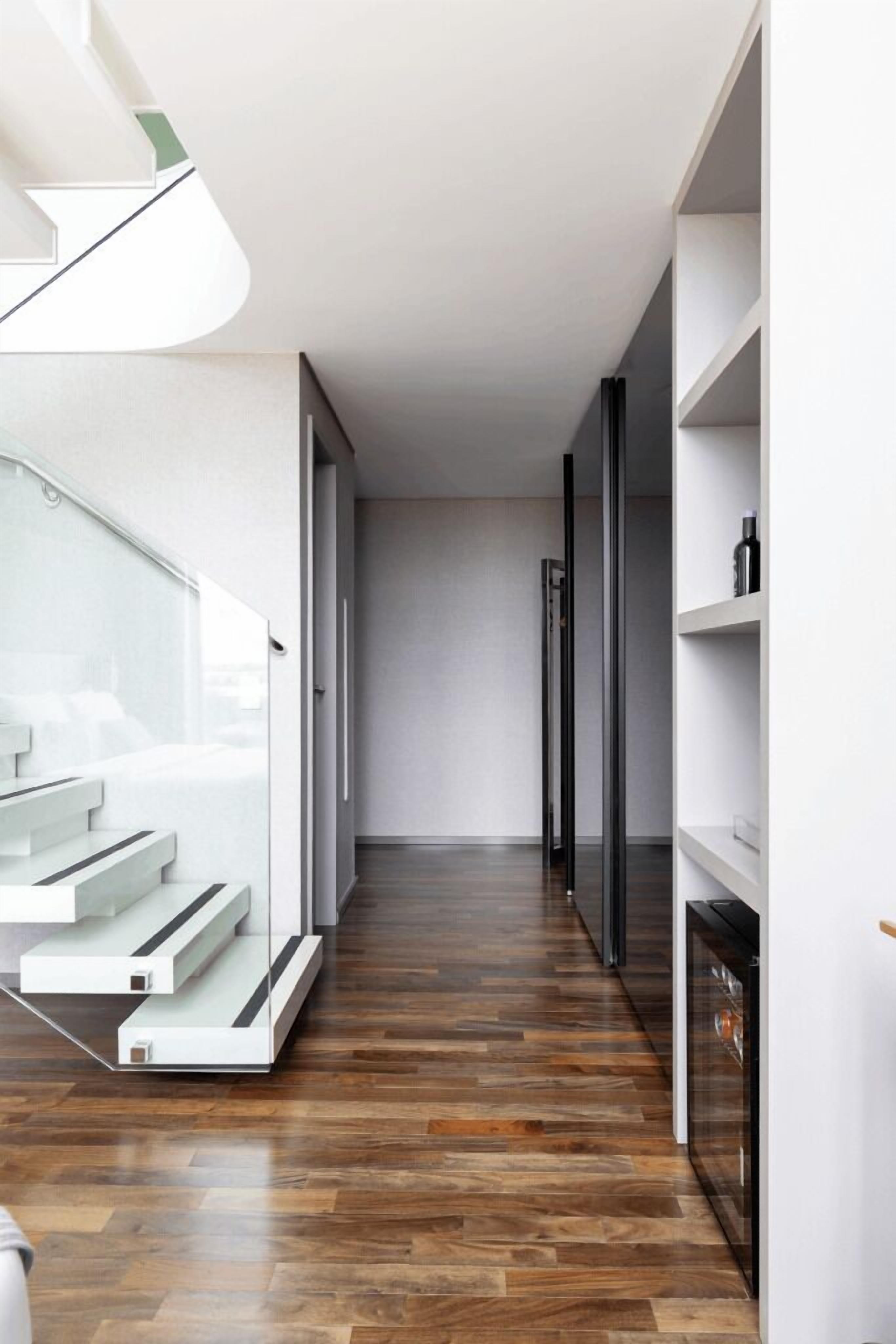 Um corredor moderno com piso de madeira, guarda-corpo de vidro e escada que leva a um andar superior.
