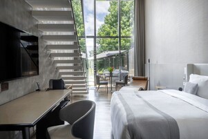 Una habitación de hotel moderna con una cama grande, un escritorio, una silla, un televisor y una escalera que lleva a un balcón con vistas a árboles.