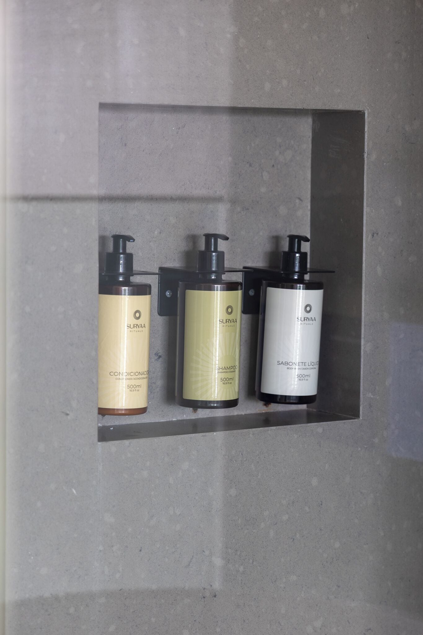 Three bottles of Surya shampoo and conditioner in a grey, tiled shower enclosure.
