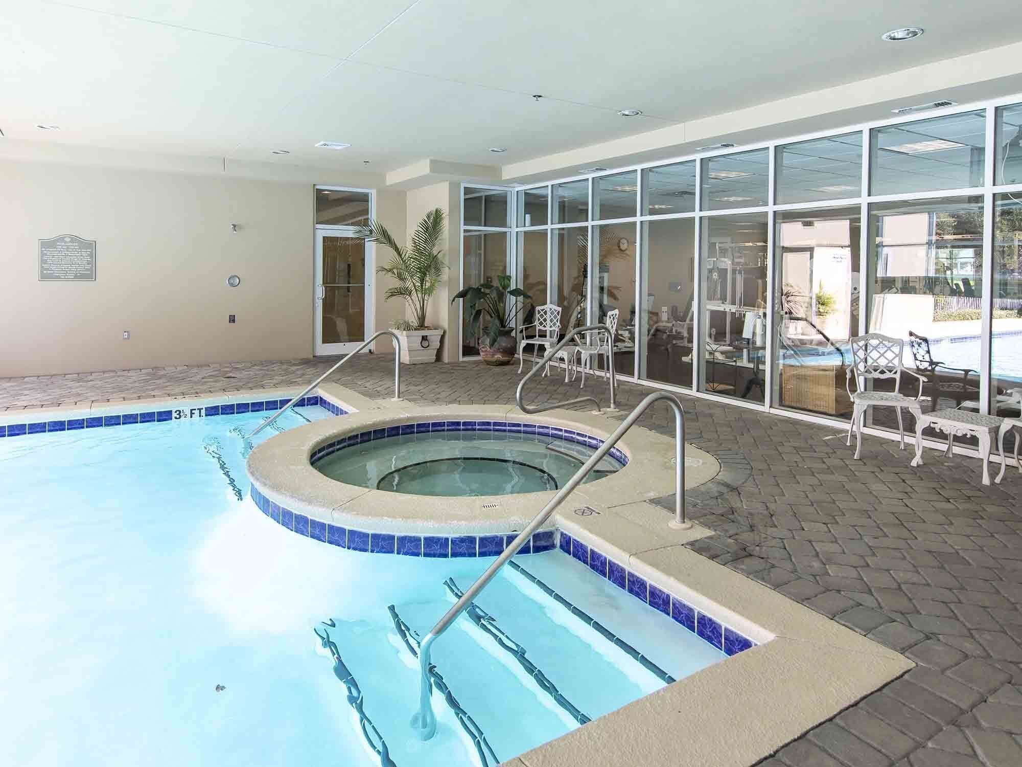 An indoor swimming pool with a hot tub, surrounded by glass doors and potted plants.