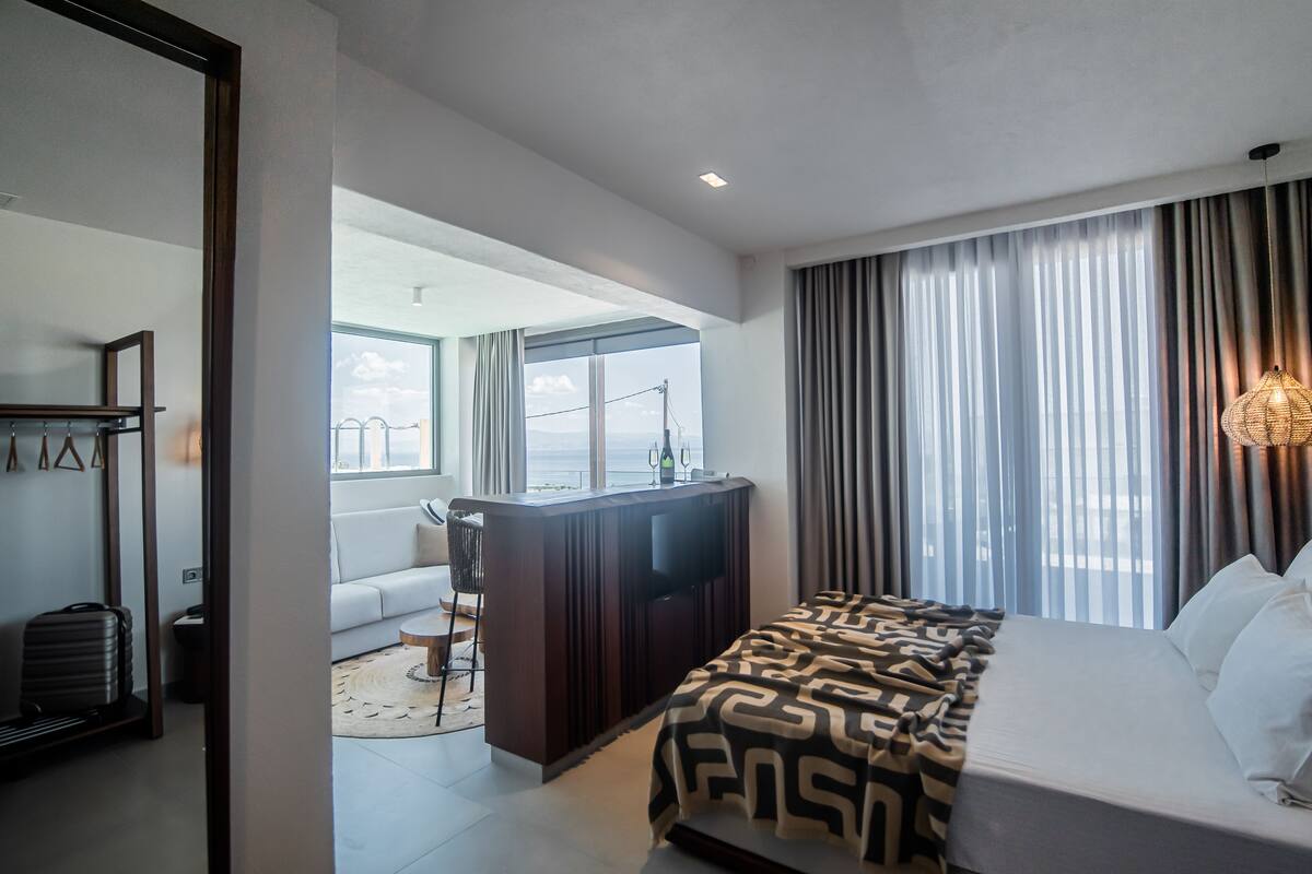 A modern hotel room with a large bed, a desk with a TV, and a view of the city through large windows.