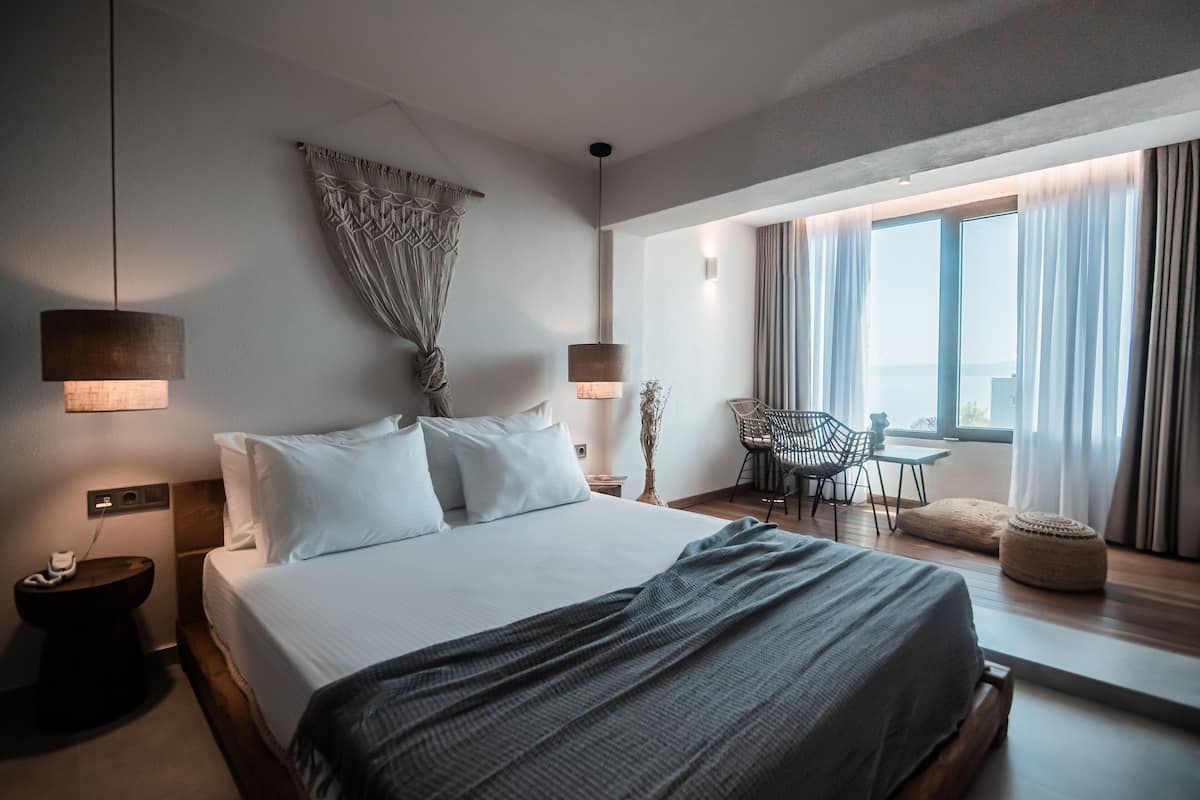 A modern hotel room with a large bed, bedside lamps, a small table with chairs, and a view of the outdoors through large windows.