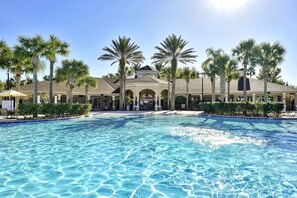 A swimming pool with a clear blue water surface, surrounded by palm trees and a building with a covered patio area. - Exquisite Escape At Windsor Hills with Pool (Kissimmee)