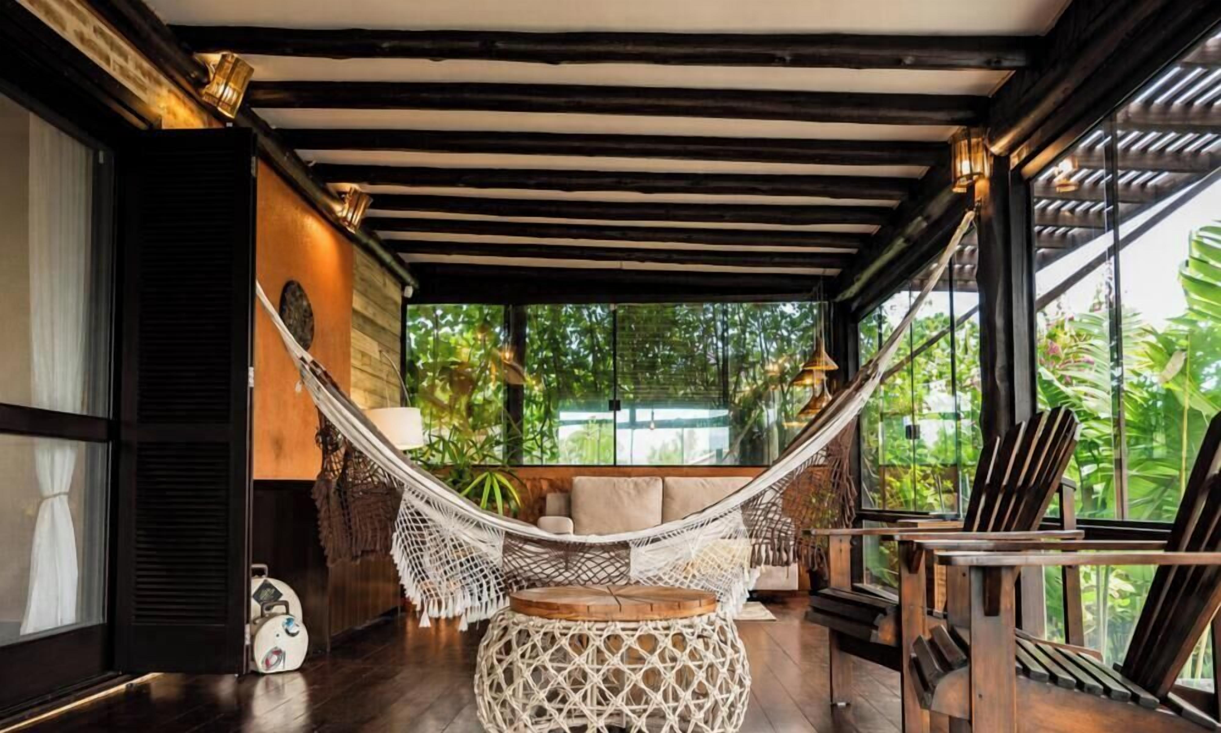 Una silla hamaca en una habitación con piso de madera, una mesa redonda tejida y vista a zonas verdes al exterior.