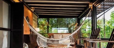 Una silla hamaca en una habitación con piso de madera, una mesa redonda tejida y vista a zonas verdes al exterior.