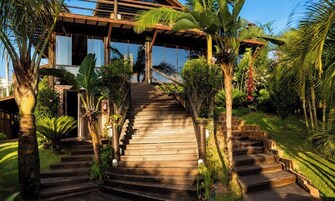 A wooden staircase leading to a modern house with a glass facade, surrounded by lush tropical vegetation.