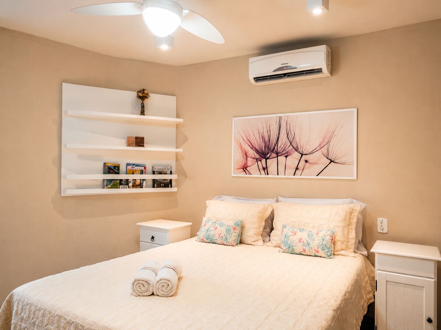 Une chambre à coucher comprenant un lit, une table de chevet, une étagère avec des livres, un climatiseur fixé au mur et un ventilateur de plafond.