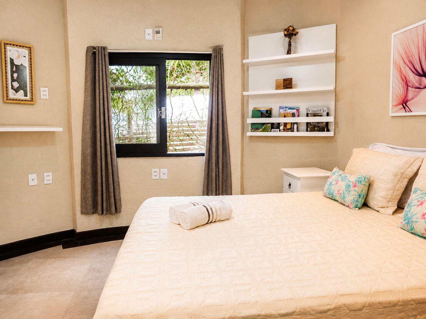 Quarto de hotel com cama, janela com cortinas, mesa de cabeceira e prateleira com livros e um vaso.