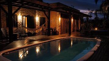A pool area with a wooden deck, a hammock, and a brick building with a covered patio.