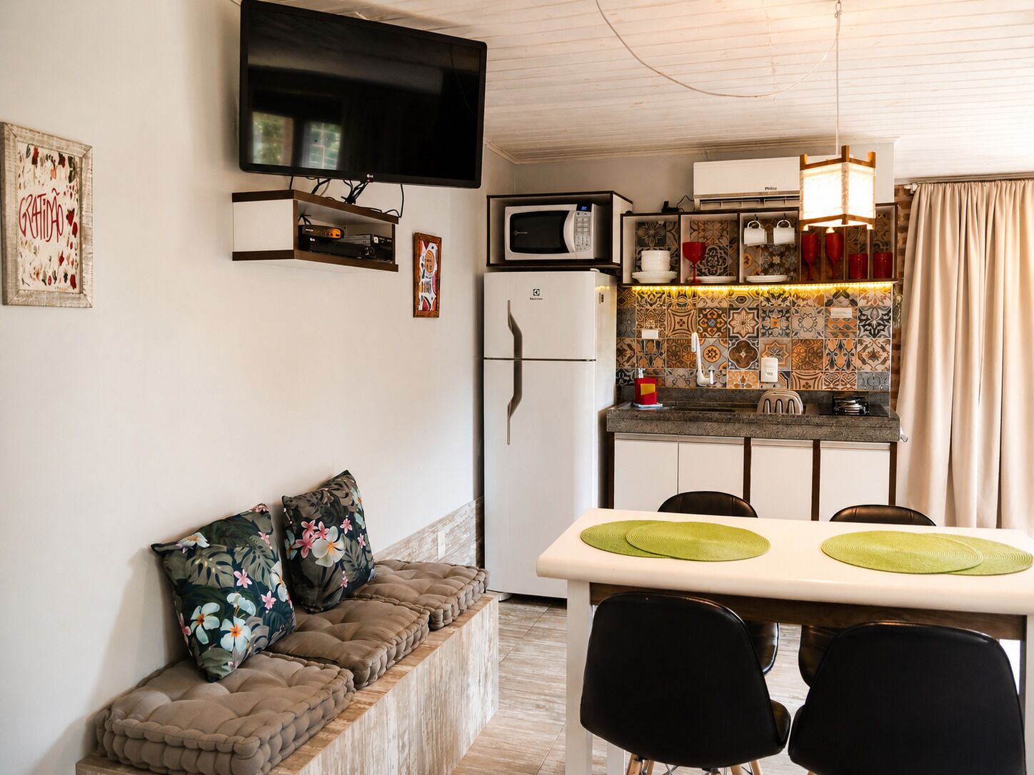 Un espacio habitable compacto con sofá, una mesa de comedor con sillas, zona de cocina con microondas y un televisor de pantalla plana sobre el sofá.