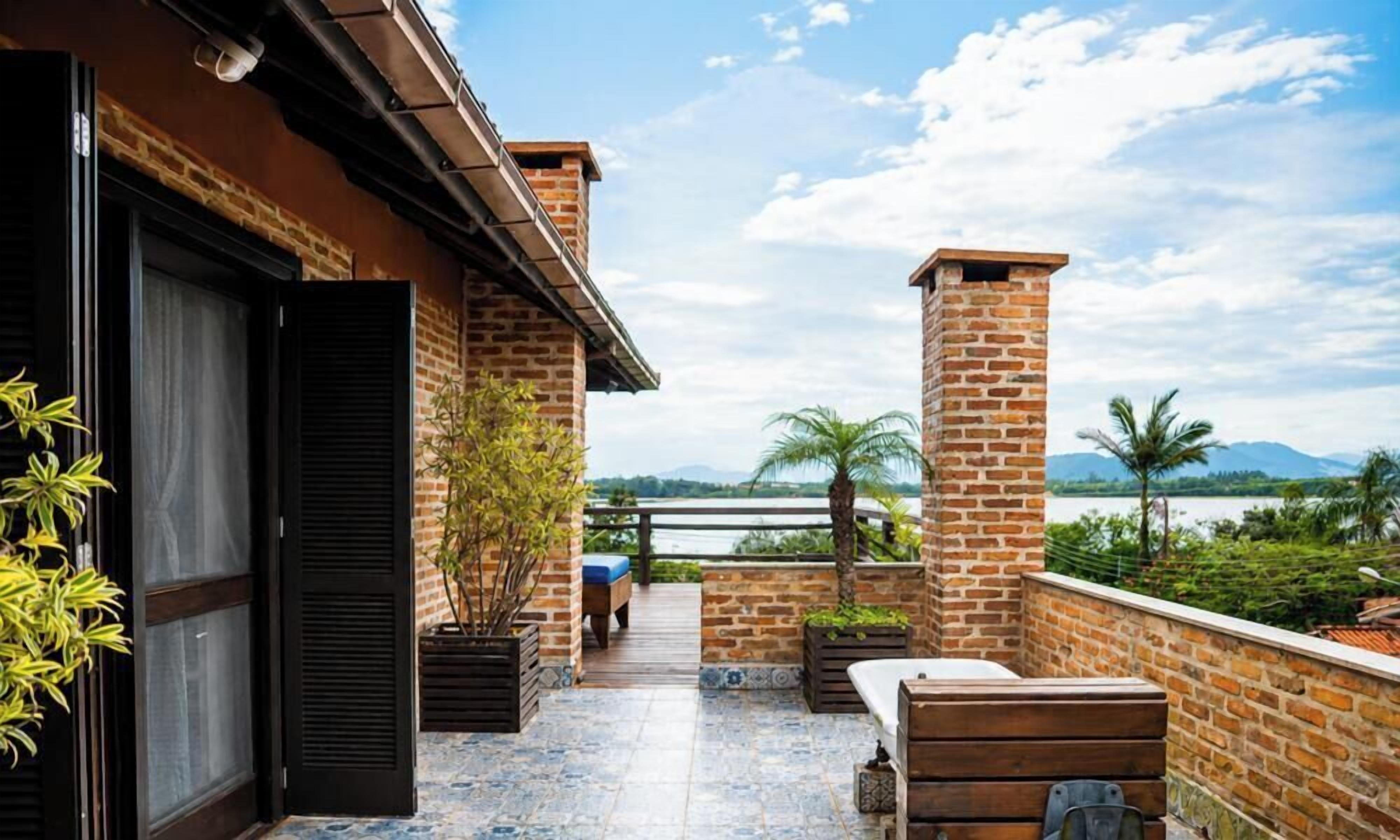 A balcony with a view of a river and mountains, a brick wall, and a wooden bench.