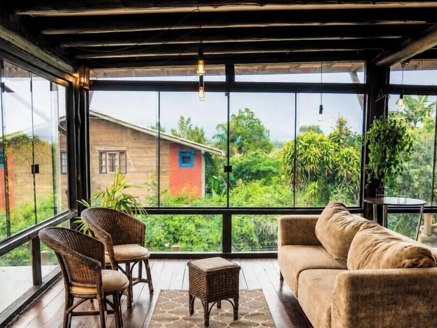 A room with a large window, wicker furniture, and a view of greenery and a building outside.
