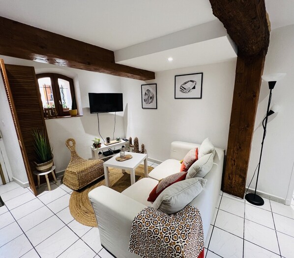 Een moderne woonkamer met een witte bank, een salontafel en een aan de muur bevestigde televisie. In de hoek staat een potplant en een rieten stoel.