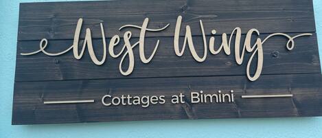 A wooden sign with the text "West Wing Cottages at Bimini" against a light blue wall.