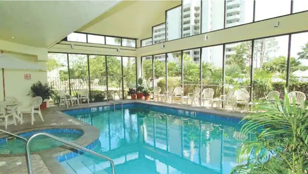 An indoor swimming pool with clear glass walls, surrounded by white chairs and potted plants. - Universal/Pool view +FREE parking. Newly renovated 2bdrm/2bth (Orlando)