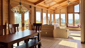 Um quarto amplo com paredes e teto de madeira, grandes janelas com vista para o exterior, uma mesa de jantar com cadeiras e um espaço de estar com sofá e almofadas.