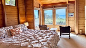 Quarto de cabana de madeira com uma cama grande, uma cadeira, uma mesa pequena e vista para o oceano através de janelas amplas.