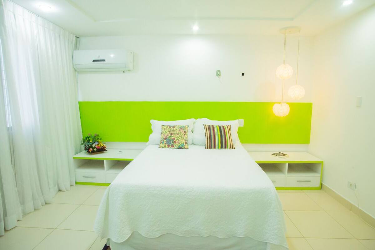 A modern hotel room with a large bed, a green accent wall, a white air conditioning unit, and a bedside table with a lamp.