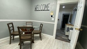 Un comedor con un cartel de 'WELCOME', una mesa de vidrio, cuatro sillas de madera y una cocina visible a través de una puerta abierta.