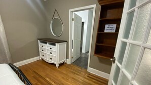 Un dormitorio con piso de madera, un tocador blanco, un espejo redondo y una puerta que da a otra habitación.