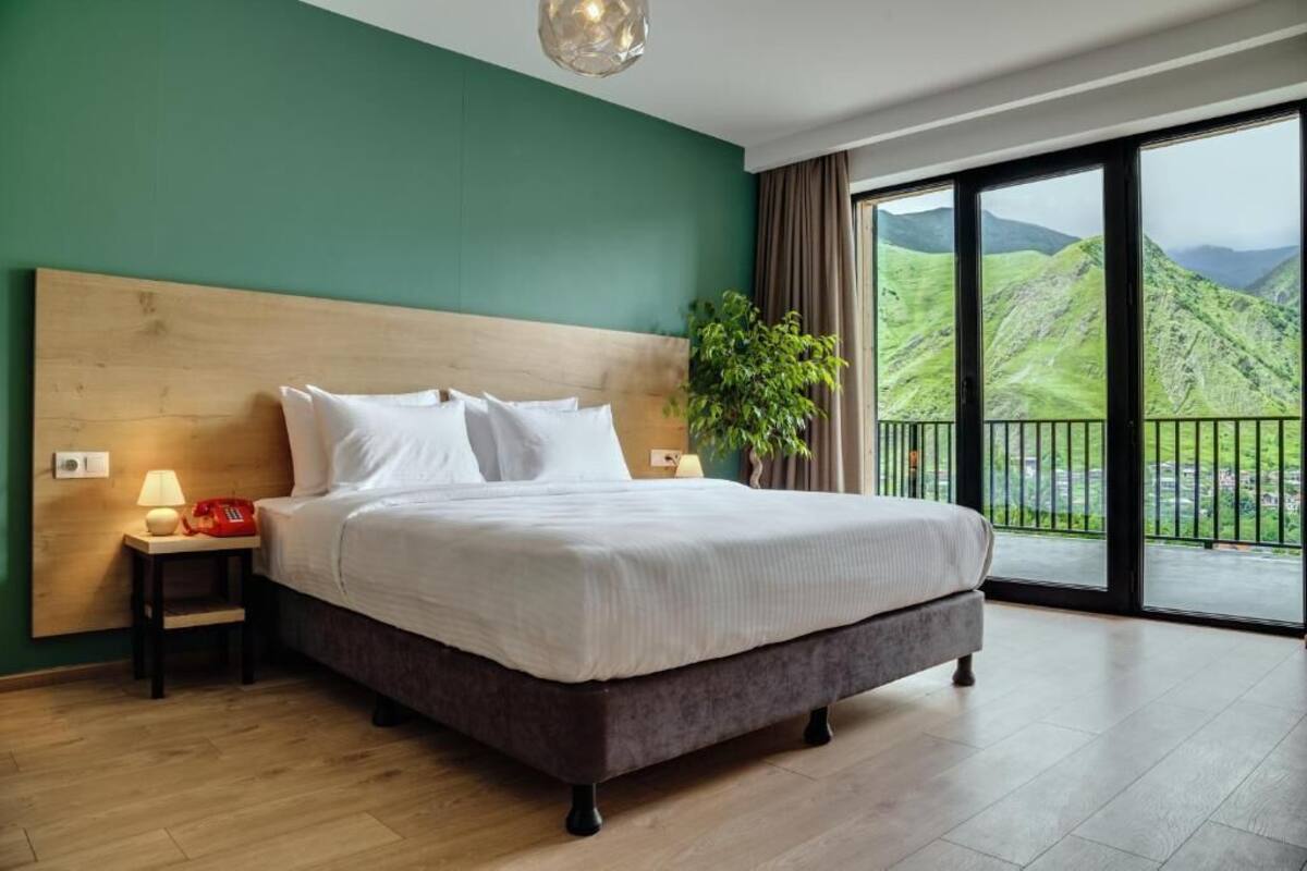 A bedroom with a large bed, a nightstand, a lamp, a plant, and a view of green hills through a balcony.