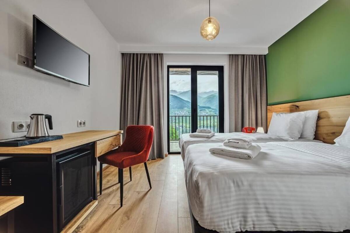 A hotel room with a bed, a desk with a kettle, a red chair, a TV mounted on the wall, and a view of mountains through the window.