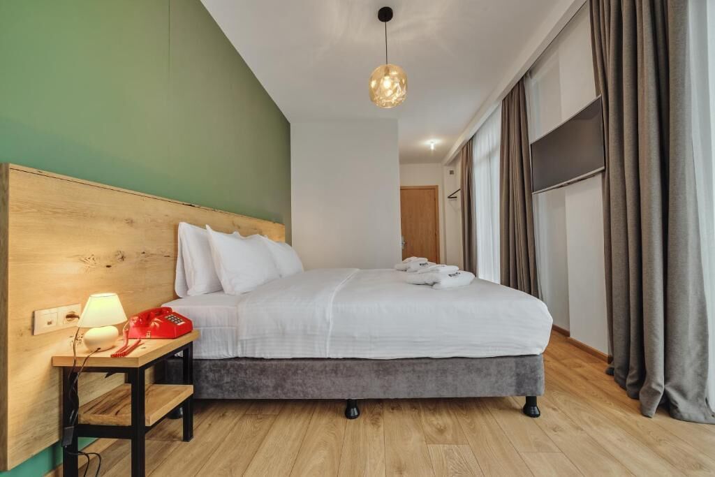 Habitación de hotel con una cama, una mesita de noche con lámpara y un teléfono rojo, una lámpara colgante y vistas al exterior a través de amplias ventanas con cortinas.