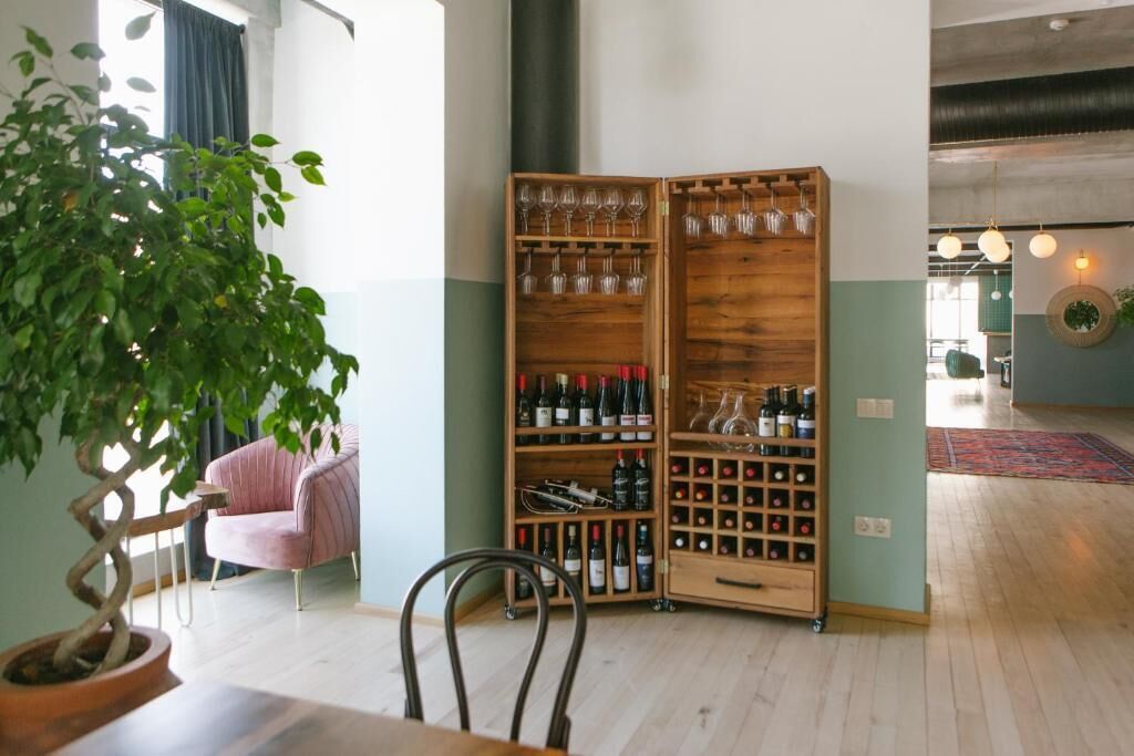 A room with a wooden wine rack holding wine bottles and glasses, a potted plant, and a pink chair.
