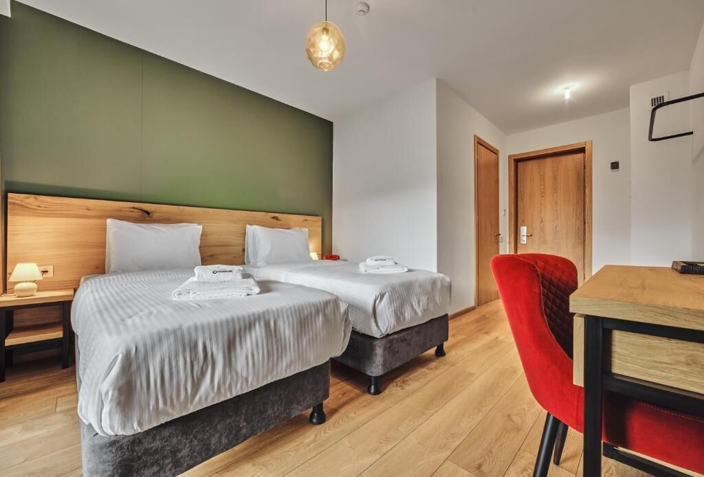 A hotel room with two beds, a wooden headboard, a red chair, and a wooden table.