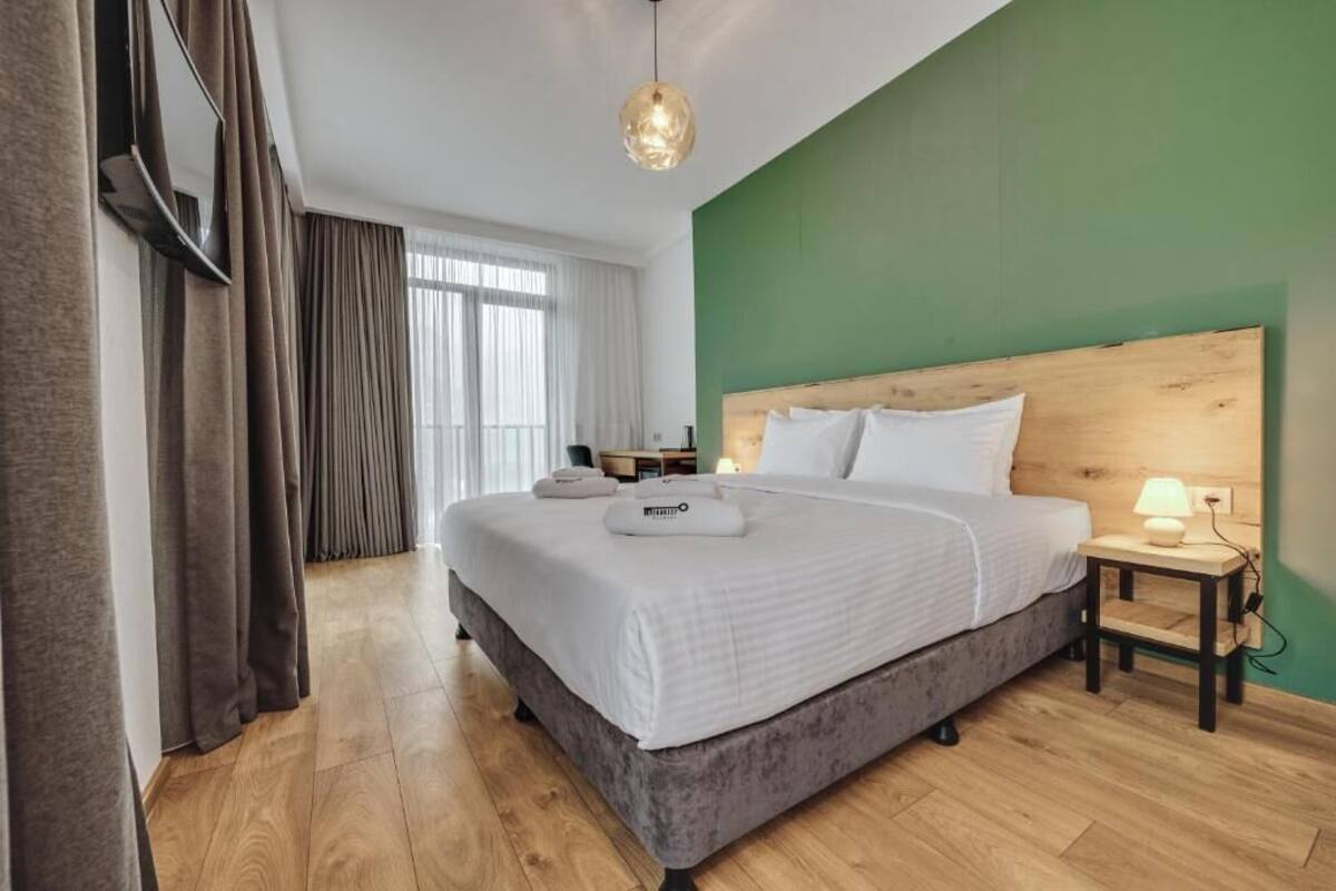 A hotel room with a large bed, a wooden headboard, a bedside table with a lamp, and a view of the street through the window.