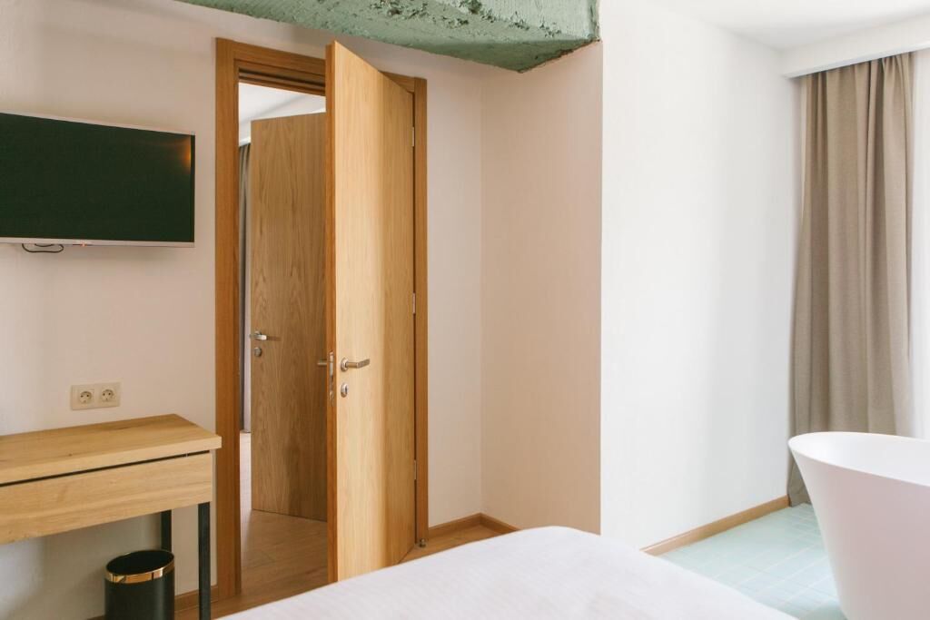 A hotel room with a wooden desk, a bed, a white bathtub, and a television mounted on the wall.