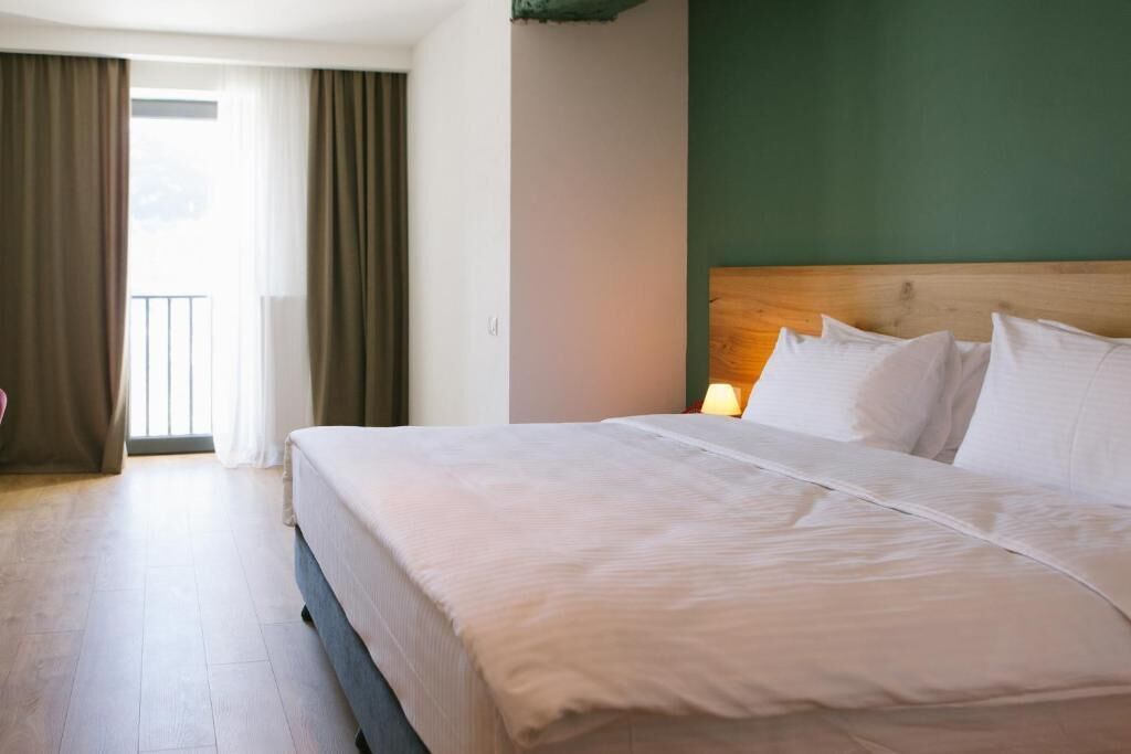 A hotel room with a large bed, a wooden headboard, and a view of the outdoors through a window with curtains.