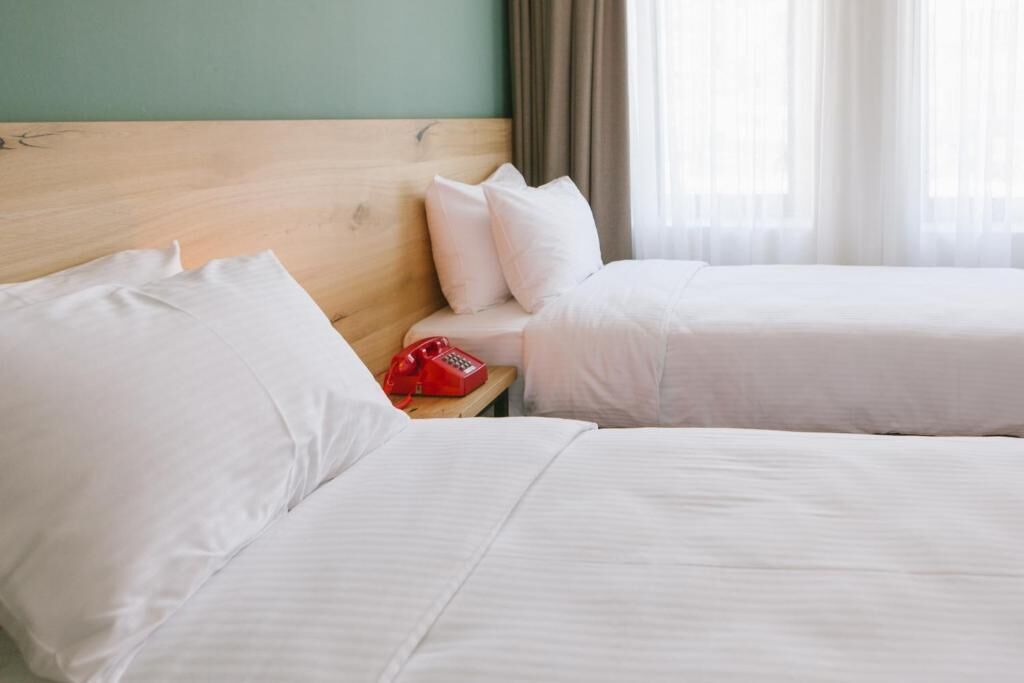 Habitación de hotel con dos camas, cabecero de madera, ropa de cama blanca, teléfono rojo y una ventana con cortinas traslúcidas.