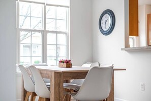 Un comedor con una mesa de madera y sillas blancas, un reloj en la pared y una ventana con vistas a árboles.