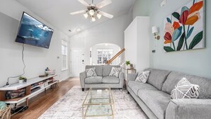 A modern living room with a grey sofa, a glass coffee table, a flat-screen TV, and a ceiling fan.