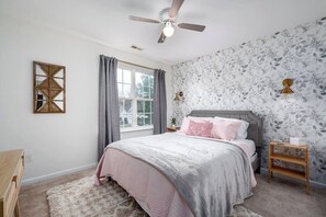 Un dormitorio con cama, ventilador de techo, ventana con cortinas, lámpara de pared y un espejo decorativo.