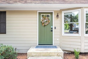 A house with a green door, a welcome mat, and a window with a view of trees.