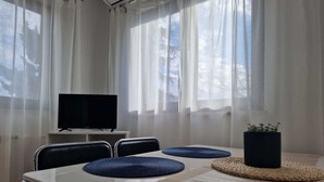 Ett hotellrum med ett matbord, stolar, en tv och tunna gardiner.