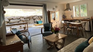 Ampla sala de estar com mesa de jantar, cadeiras, sofá e mesa de centro. Há uma área de cozinha com pia e armários. O ambiente possui uma grande janela que oferece luz natural e vista para o exterior.
