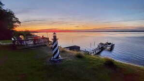 Ein Leuchtturm, eine Anlegestelle und eine Terrasse mit Stühlen mit Blick auf ein Gewässer bei Sonnenuntergang.